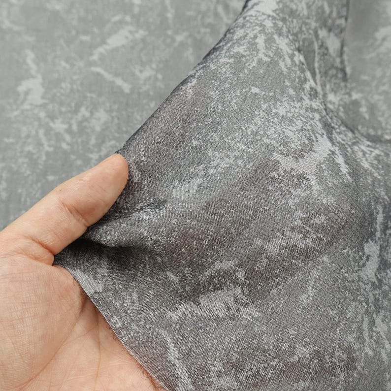 May include: Close-up of a gray fabric with a textured, abstract pattern. The fabric is held up, revealing its semi-sheer quality. The pattern features lighter gray and white accents against a darker gray background, creating a mottled effect. The fabric appears soft and flowing.