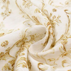 May include: Close-up of a cream-colored fabric with a repeating pattern of gold-colored floral and leaf designs. The fabric is draped, showing folds and texture. The pattern is detailed and elegant.