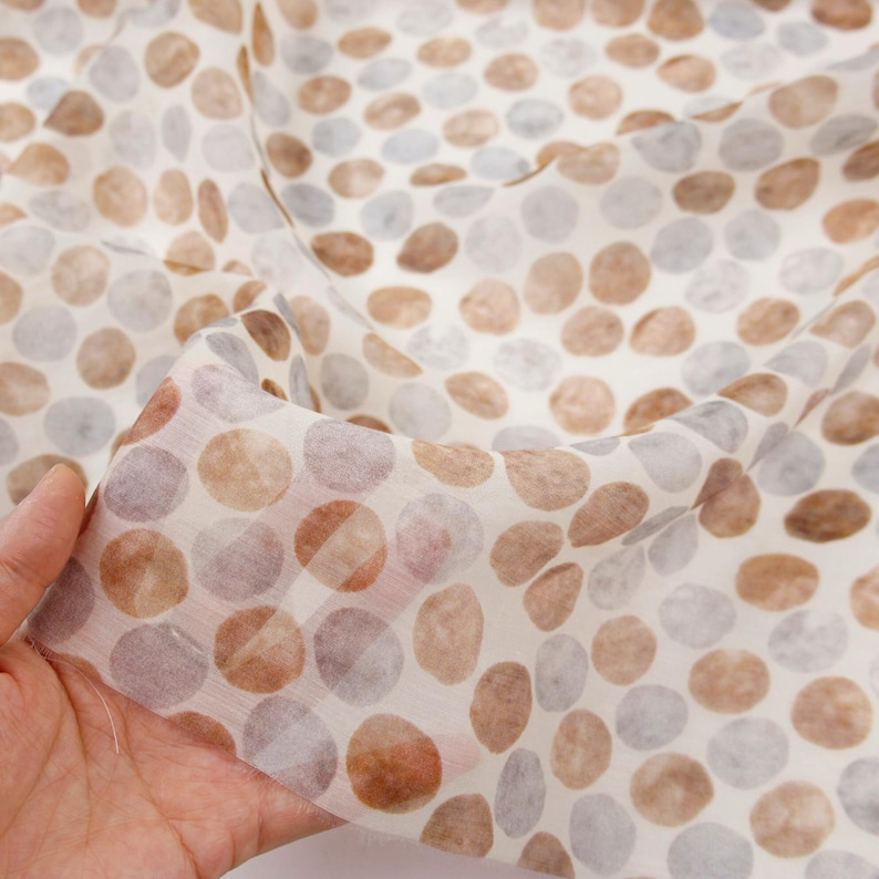 May include: A sheer fabric with a pattern of brown and gray circles on a white background. The fabric is held up, showing its delicate, translucent quality. The circles vary in size and shade.
