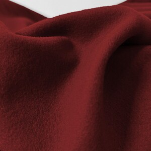 Puede incluir: Primer plano de una tela rojo oscuro con una superficie suave y texturizada. El material está drapeado, creando pliegues y sombras que resaltan su apariencia afelpada. El color es un tono rojo rico y saturado.