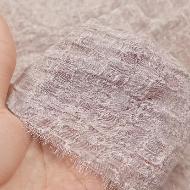 May include: Close-up of a piece of light purple fabric with a textured, geometric pattern. The fabric has a delicate, sheer appearance and a fringed edge. The fabric is held in a hand.