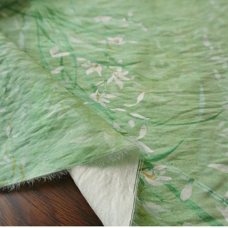 Peut inclure: Tissu vert clair avec un motif floral de fleurs blanches et de tiges vertes. Le tissu pr&eacute;sente une texture l&eacute;g&egrave;rement froiss&eacute;e, avec un motif de style aquarelle. Le tissu est pli&eacute;, r&eacute;v&eacute;lant un envers blanc.