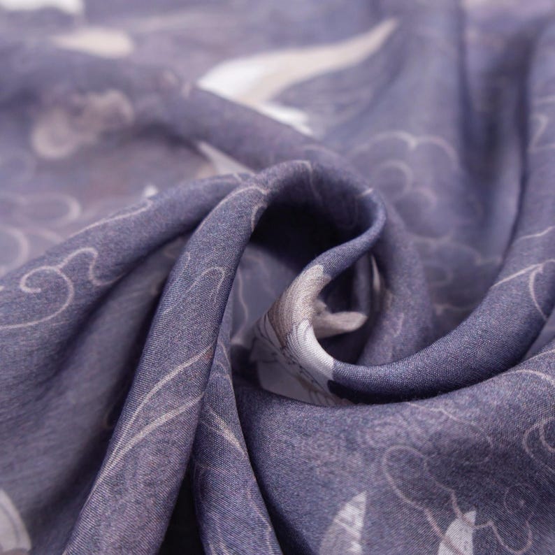 May include: Close-up of a folded fabric with a repeating pattern of white birds and cloud-like shapes on a muted purple background. The fabric has a soft, flowing texture, with subtle variations in color and tone.