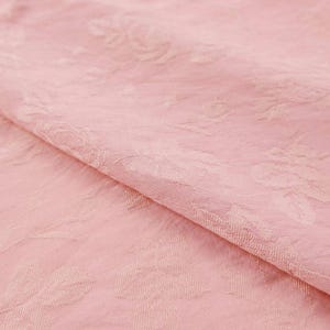 May include: A light pink fabric featuring a tonal jacquard weave with a repeating floral pattern of roses and leaves. The material has a soft, slightly textured surface with a subtle sheen, showcasing the intricate woven design across the textile.