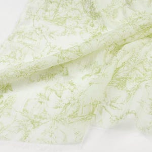 May include: A white fabric with a light green toile pattern. The pattern features floral and architectural elements. The fabric is draped, showing its texture and design. The overall aesthetic is delicate and classic.