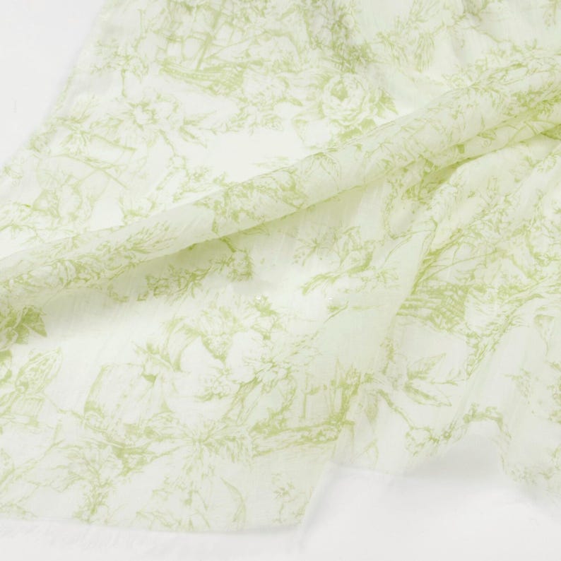 May include: A white fabric with a light green toile pattern. The pattern features floral and architectural elements. The fabric is draped, showing its texture and design. The overall aesthetic is delicate and classic.