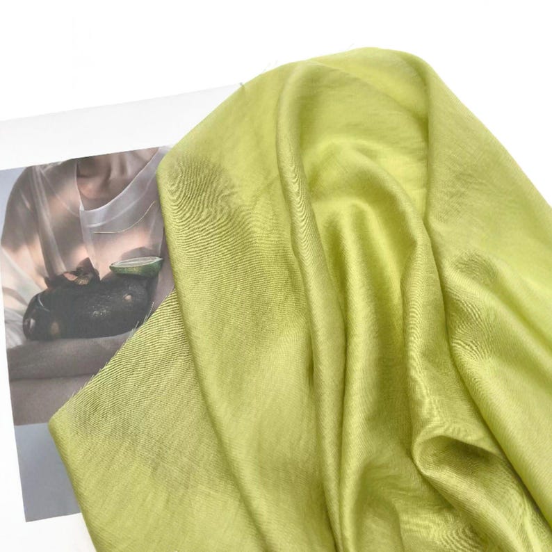 May include: A light lime green scarf with a subtle, fine-textured weave. The fabric displays a soft, luminous sheen and drapes in loose, flowing folds. The material appears lightweight and semi-translucent, showing a smooth, consistent color.