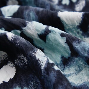 May include: Close-up of a fabric with a watercolor-like pattern. The fabric features a blend of dark navy, light blue, and white splotches, creating a flowing, abstract design. The material appears soft and slightly wrinkled.