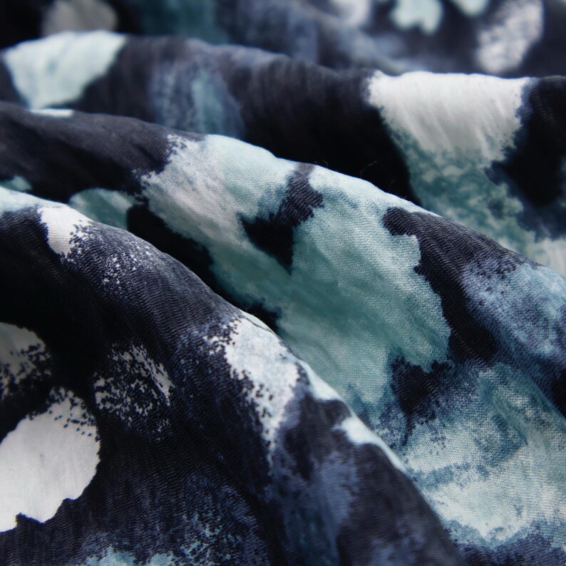 May include: Close-up of a fabric with a watercolor-like pattern. The fabric features a blend of dark navy, light blue, and white splotches, creating a flowing, abstract design. The material appears soft and slightly wrinkled.