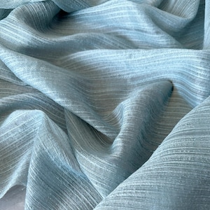 Puede incluir: Una tela de color azul claro con una textura acanalada horizontal distintiva. El material está drapeado en pliegues suaves y fluidos, creando una serie de sombras y luces que resaltan el patrón lineal en la superficie del tejido.