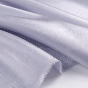 May include: Close-up of a light lavender fabric with a subtle sheen. The fabric is draped, showing soft folds and a textured surface. The edges are unfinished, revealing a frayed texture. The material appears lightweight and flowing.