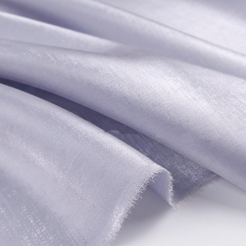 May include: Close-up of a light lavender fabric with a subtle sheen. The fabric is draped, showing soft folds and a textured surface. The edges are unfinished, revealing a frayed texture. The material appears lightweight and flowing.