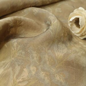 May include: Close-up of a beige fabric with a subtle floral pattern. The fabric has a soft sheen and is draped in gentle folds. A cream-colored flower rests on the fabric, adding a touch of elegance. The fabric's texture is visible, suggesting a luxurious material.