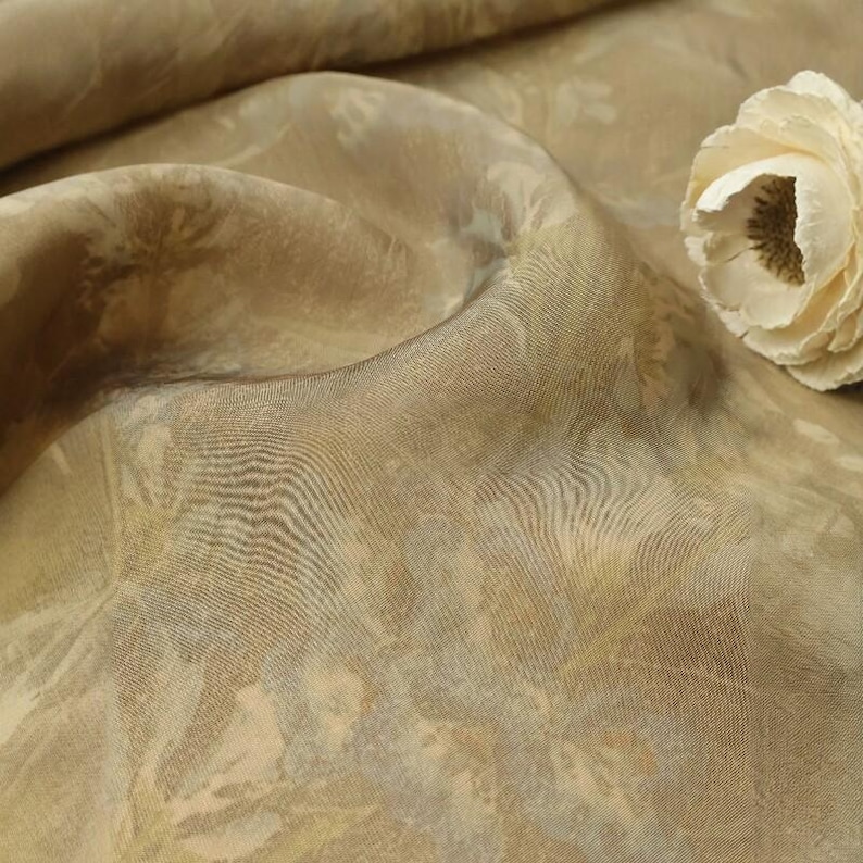 May include: Close-up of a beige fabric with a subtle floral pattern. The fabric has a soft sheen and is draped in gentle folds. A cream-colored flower rests on the fabric, adding a touch of elegance. The fabric's texture is visible, suggesting a luxurious material.