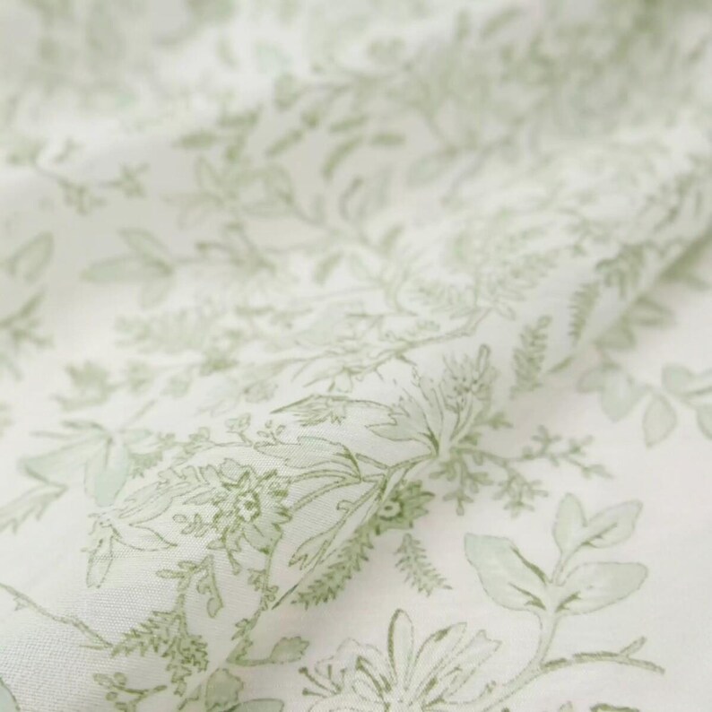 Puede incluir: Primer plano de una tela blanca con un delicado estampado floral en tonos verdes. El dise&ntilde;o presenta ilustraciones bot&aacute;nicas detalladas de hojas y flores, creando una est&eacute;tica suave y natural. La tela parece ligera y fluida.