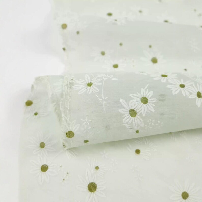 May include: A sheer, light green fabric featuring a repeating pattern of white daisy flowers with olive green centers. The material is translucent and lightweight, displaying a delicate floral print across the entire surface of the textile.