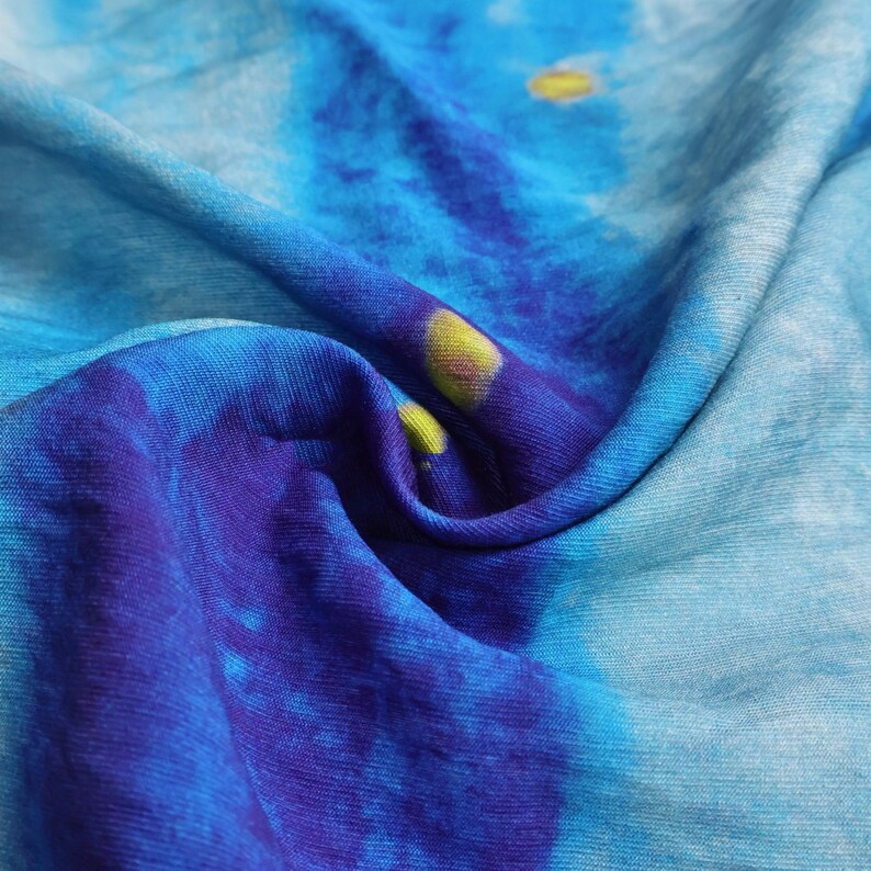May include: Close-up of a fabric with a tie-dye pattern in shades of blue and a touch of yellow. The fabric is soft and appears to be draped, with visible folds and textures. The design has a watercolor effect, with the colors blending seamlessly.