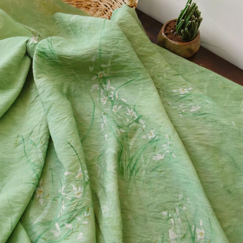 Peut inclure: Tissu vert clair avec un motif floral d&eacute;licat. Le tissu pr&eacute;sente un motif de fleurs blanches et de tiges vertes, cr&eacute;ant une esth&eacute;tique douce et naturelle. Le tissu est drap&eacute;, r&eacute;v&eacute;lant sa texture et son mouvement.