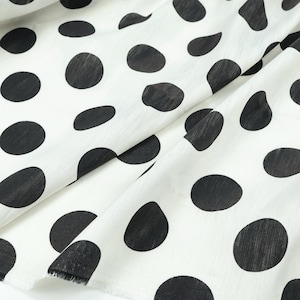 May include: White fabric with large, irregular black polka dots. The fabric is folded, showing the texture and pattern. The dots vary in size and shape, creating a playful and classic design. The fabric's edges are visible.