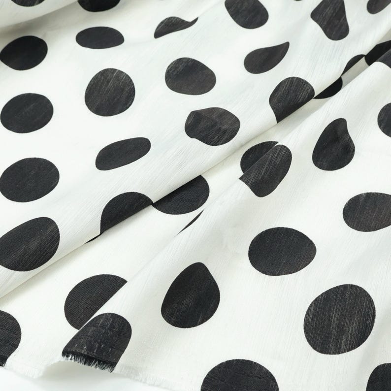 May include: White fabric with large, irregular black polka dots. The fabric is folded, showing the texture and pattern. The dots vary in size and shape, creating a playful and classic design. The fabric's edges are visible.