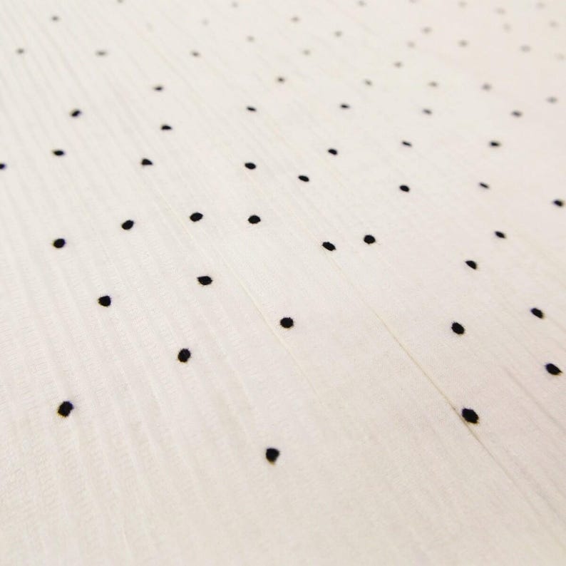 May include: Close-up of a white fabric with a subtle ribbed texture, adorned with evenly spaced black polka dots. The dots are small and create a simple, yet elegant pattern. The fabric appears soft and lightweight.