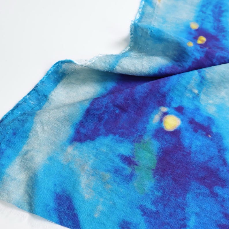 May include: A close-up of a blue and white fabric with a watercolor effect. The fabric has a frayed edge and features shades of blue, with splashes of yellow. The overall design is abstract and artistic.