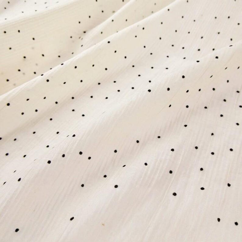 May include: Close-up of a white fabric with a subtle ribbed texture. Scattered across the surface are small, evenly spaced black polka dots. The fabric is gently folded, creating soft shadows and highlighting the texture.