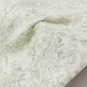 Puede incluir: Primer plano de una tela blanca con un delicado estampado de hojas verdes y elementos florales. La tela est&aacute; drapeada, mostrando pliegues y texturas suaves. El dise&ntilde;o presenta varias formas de hojas y peque&ntilde;as flores.
