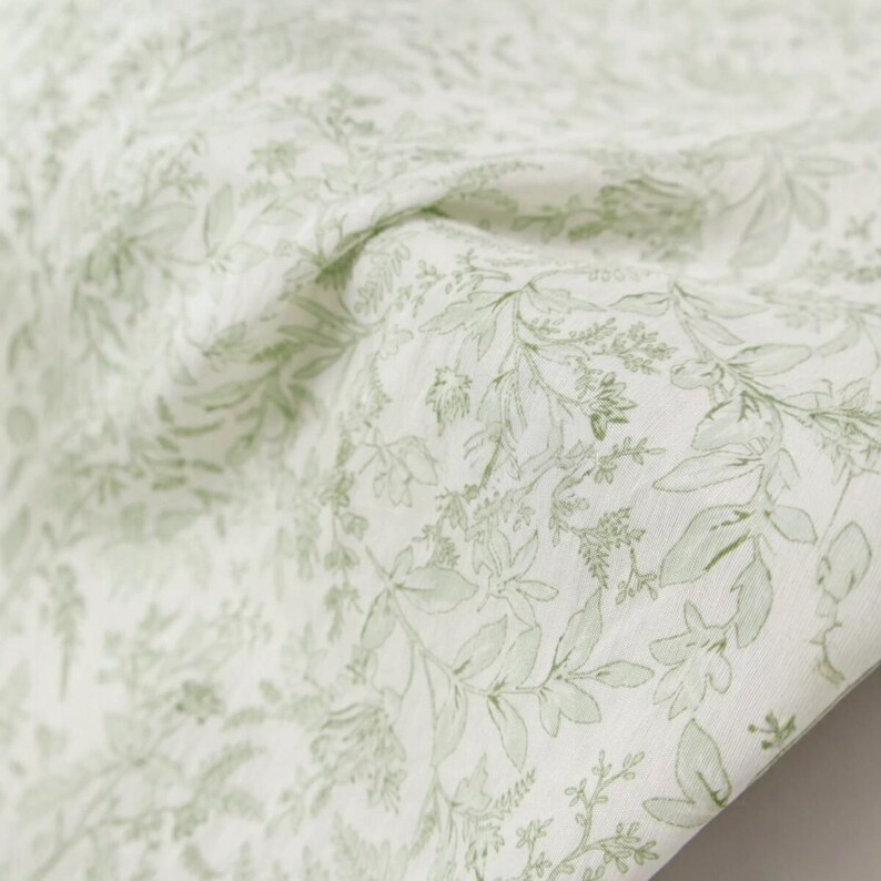 Puede incluir: Primer plano de una tela blanca con un delicado estampado de hojas verdes y elementos florales. La tela est&aacute; drapeada, mostrando pliegues y texturas suaves. El dise&ntilde;o presenta varias formas de hojas y peque&ntilde;as flores.