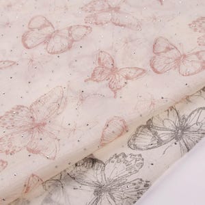 Op de afbeelding: Close-up van een stof met een herhalend vlinderpatroon. De vlinders zijn in roze en grijstinten, met fijne details. De stof heeft een lichte achtergrond en is besprenkeld met kleine, glanzende accenten.
