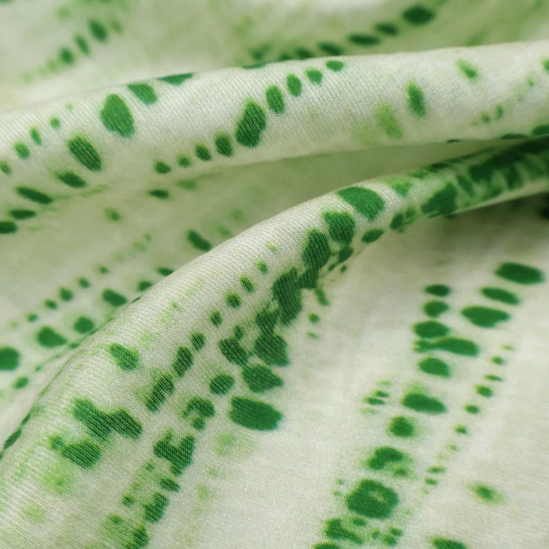 Op de afbeelding: Een zachte stof met een witte basiskleur en een herhalend patroon van levendige groene stippen gerangschikt in diagonale lijnen. Het materiaal is gevouwen om zachte, vloeiende rondingen te cre&euml;ren die de textuur en het stippenpatroon benadrukken.