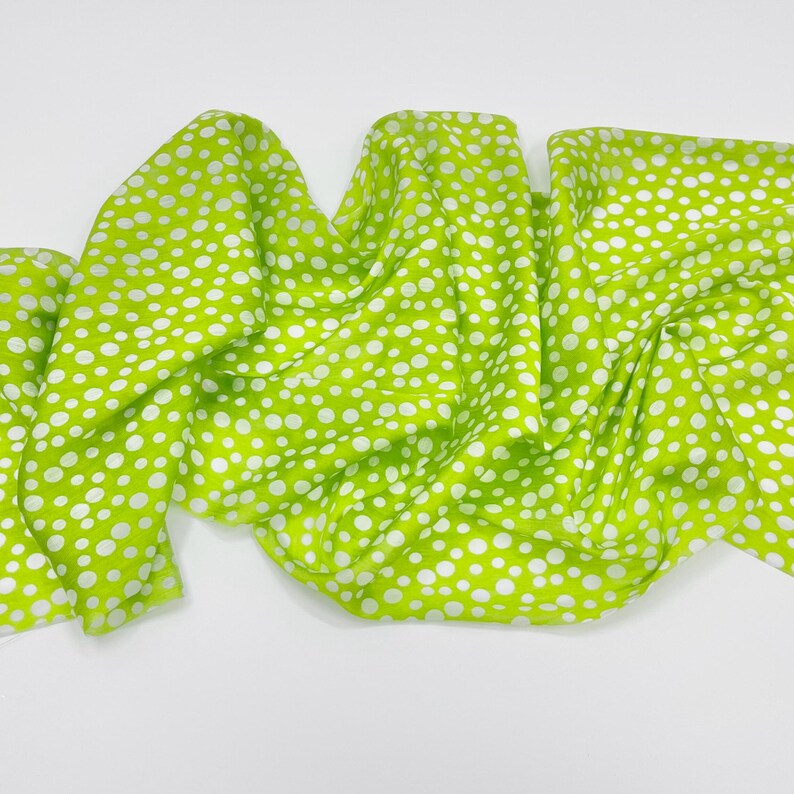 May include: A bright lime green scarf featuring a repeating pattern of white polka dots of varying sizes. The fabric is draped in soft, flowing folds, creating a textured appearance across the surface of the lightweight material.