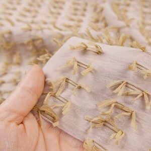 May include: A light beige fabric featuring a textured surface with small, evenly spaced tufted fringe details in a darker tan shade. The material is semi-sheer and lightweight, displaying a repeating pattern of these short, frayed thread accents.