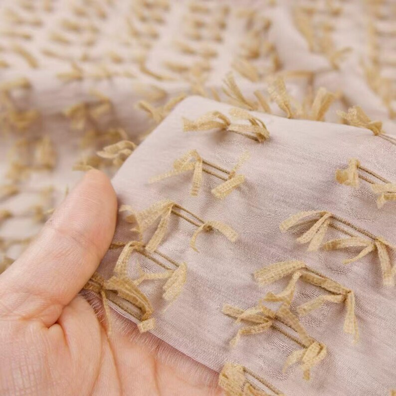 May include: A light beige fabric featuring a textured surface with small, evenly spaced tufted fringe details in a darker tan shade. The material is semi-sheer and lightweight, displaying a repeating pattern of these short, frayed thread accents.