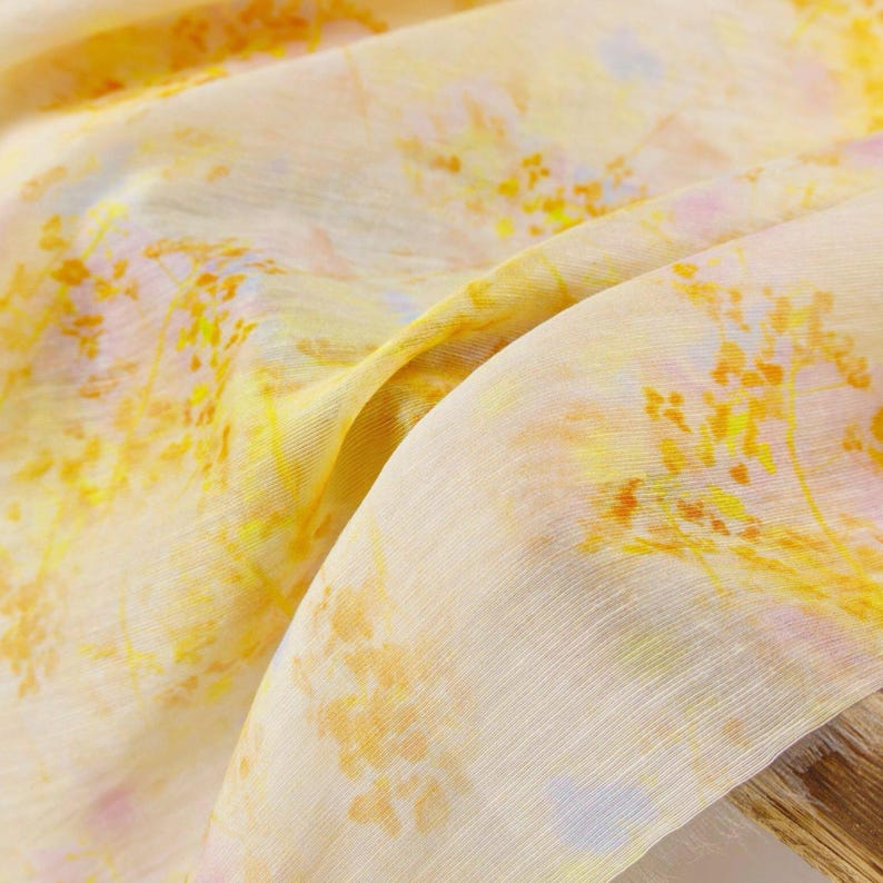 May include: A close-up of a flowing fabric with a delicate floral pattern. The fabric is primarily cream-colored, with yellow, orange, and hints of pink and blue floral accents. The material appears lightweight and slightly translucent.