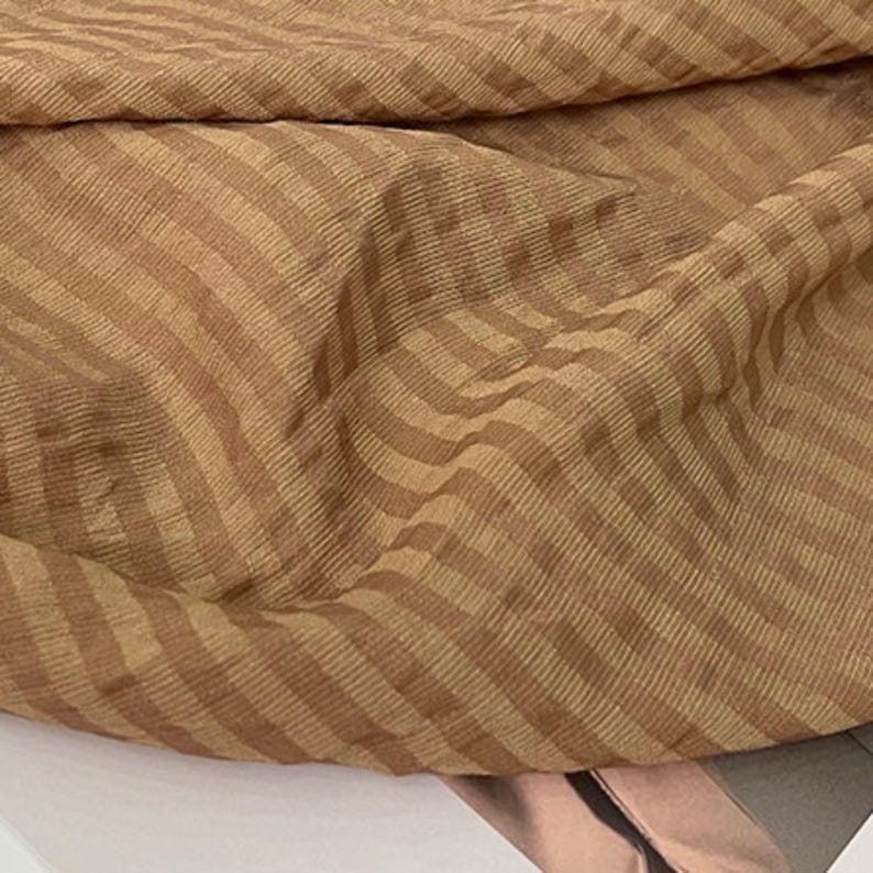 Peut inclure: Un morceau de tissu pr&eacute;sentant un motif &agrave; rayures marron clair et marron fonc&eacute;. Le mat&eacute;riau poss&egrave;de une texture c&ocirc;tel&eacute;e visible avec des lignes diagonales traversant la surface, cr&eacute;ant une profondeur et une dimension subtiles dans le textile pli&eacute;.