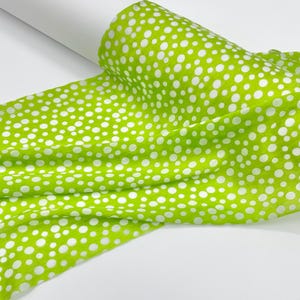 May include: A roll of fabric featuring a vibrant lime green background with a repeating pattern of white polka dots of varying sizes. The material is draped in soft folds, showcasing the consistent print across the textured textile surface.