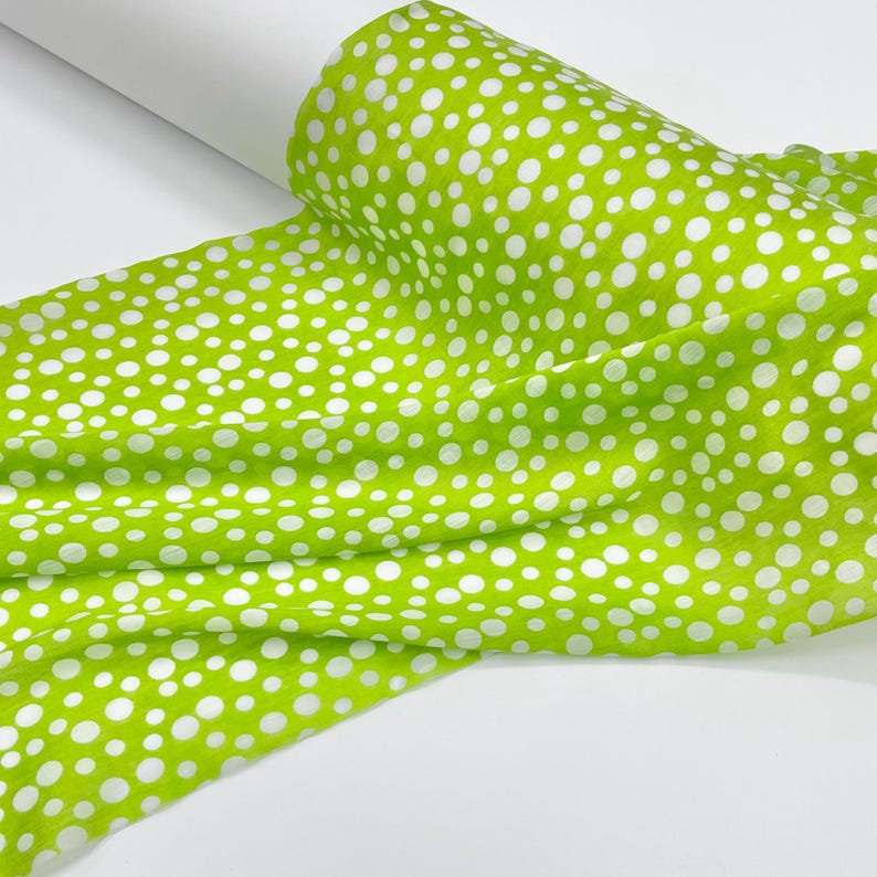 May include: A roll of fabric featuring a vibrant lime green background with a repeating pattern of white polka dots of varying sizes. The material is draped in soft folds, showcasing the consistent print across the textured textile surface.