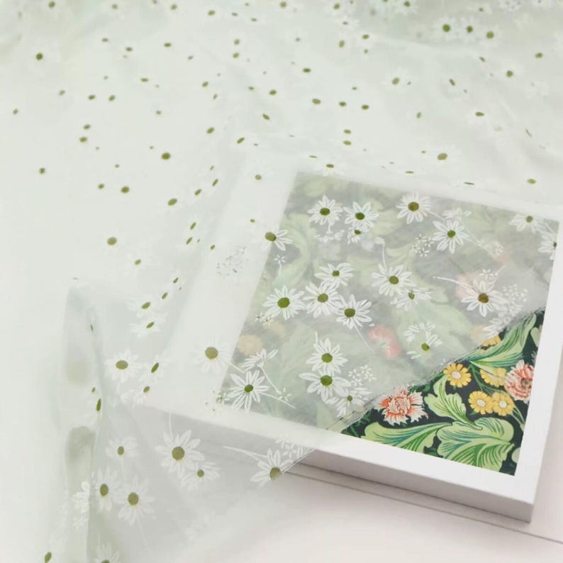 May include: A sheer white fabric featuring a repeating pattern of small white daisies with yellow centers. The fabric is draped over a square white frame that displays a vibrant botanical print with green leaves, yellow flowers, and orange blossoms.