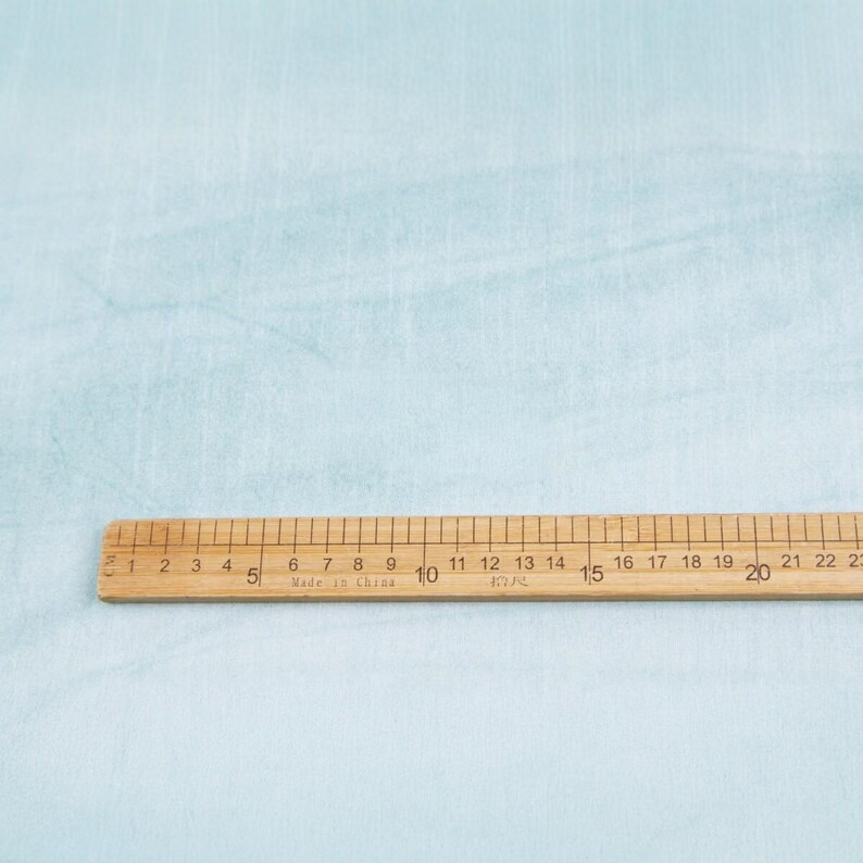 K&ouml;nnte beinhalten: Ein h&ouml;lzernes Lineal mit schwarzen Markierungen und Zahlen, das bis zu 58 cm misst, liegt auf einem hellblauen Stoff. Auf dem Lineal steht "Made in China". Der Stoff hat ein subtiles, strukturiertes Aussehen.