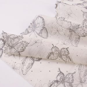 Op de afbeelding: Een cr&egrave;mekleurige sjaal met een herhalend patroon van grijze vlinderillustraties en kleine, verspreide zilveren versieringen. De sjaal is opgevouwen en toont het gedetailleerde ontwerp en de textuur van de stof.