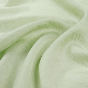 May include: Close-up of a light green fabric with a subtle, textured pattern. The fabric is draped, creating soft folds and shadows. The overall appearance is smooth and delicate, suggesting a lightweight material.