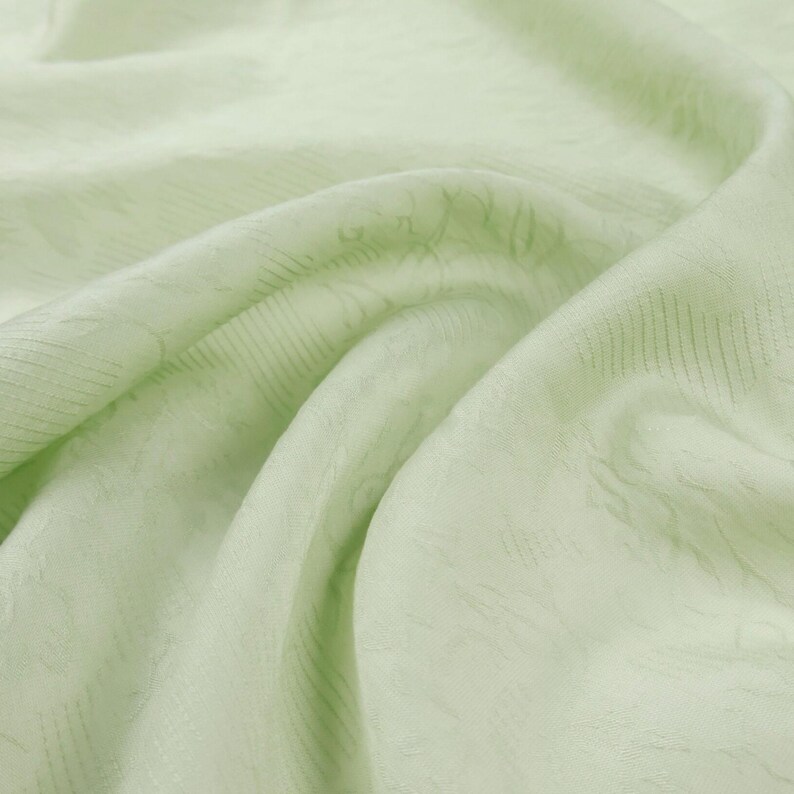 May include: Close-up of a light green fabric with a subtle, textured pattern. The fabric is draped, creating soft folds and shadows. The overall appearance is smooth and delicate, suggesting a lightweight material.