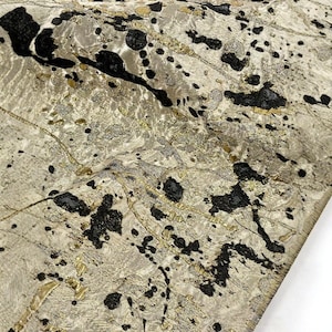 Peut inclure: Gros plan sur un tissu texturé avec une base crème. Le tissu présente des éclaboussures noires et des accents métalliques dorés et argentés. Le design a une qualité abstraite et artistique, adapté à la tapisserie ou à la mode.