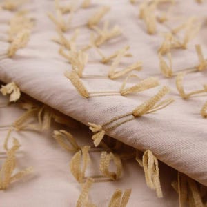 May include: A light beige fabric featuring a textured surface with numerous small, looped tufts of tan thread arranged in a repeating pattern across the material. The fabric is folded, showing the depth and the raised nature of the tufted details.