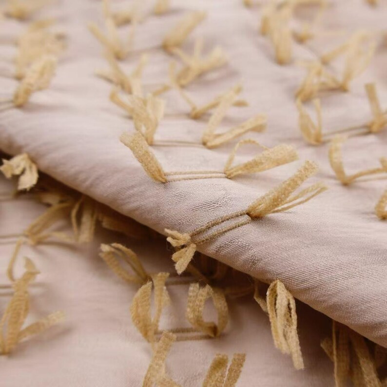 May include: A light beige fabric featuring a textured surface with numerous small, looped tufts of tan thread arranged in a repeating pattern across the material. The fabric is folded, showing the depth and the raised nature of the tufted details.
