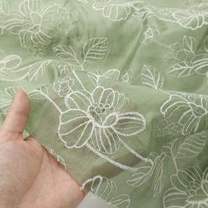 May include: A sage green fabric featuring a delicate white embroidered floral pattern with outlined petals and leaves. The material is semi-sheer and lightweight, showcasing a repeating botanical design across the entire surface of the textile.