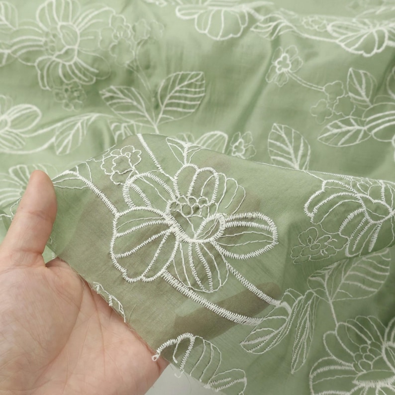 May include: A sage green fabric featuring a delicate white embroidered floral pattern with outlined petals and leaves. The material is semi-sheer and lightweight, showcasing a repeating botanical design across the entire surface of the textile.