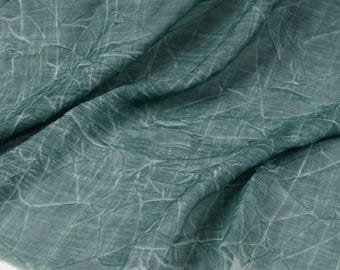 May include: A teal-colored fabric featuring a distinct crinkled texture throughout the surface. The material displays a pattern of fine, light-colored lines that crisscross across the deep blue-green base, creating a complex, web-like visual effect.