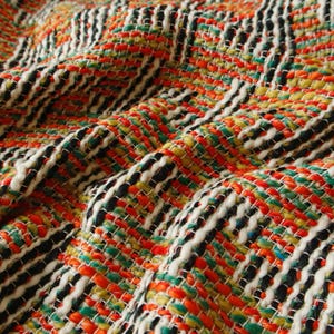 May include: Close-up of a woven fabric with a textured surface. The fabric features a colourful pattern of orange, green, yellow, black, and white threads. The weave creates a diagonal striped design, with the threads interlacing to form a textured surface.